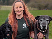 White female with long red hair wearing Jerry Green Dog Rescue green t-shirt with old black Labrador on her right and black spaniel on her left.