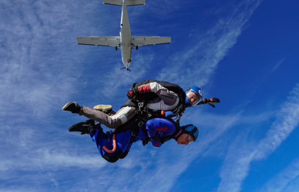 An image of someone doing a tandem skydive against bright blue skies and the plane they jumped from above.