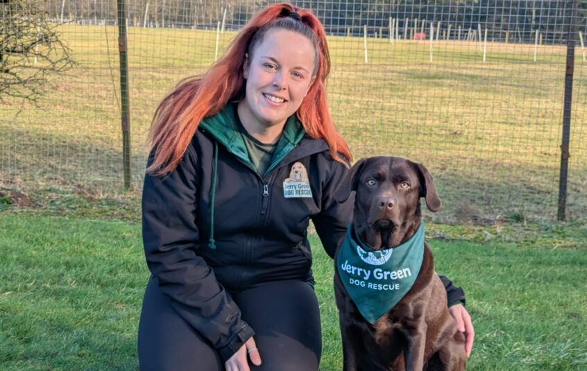 White female knelt in grassy field with Chocolate Labrador sat next to her wearing a Jerry Green Dog Rescue bandana